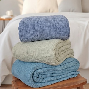Peut inclure: Une pile de trois couvertures en coton dans les tons de bleu et de vert. La couverture supérieure est roulée, bleu clair. La couverture du milieu est pliée, vert clair. La couverture inférieure est pliée, bleue. Les couvertures sont sur un tabouret en bois.