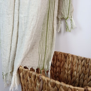 May include: A white linen throw with green stripes and tassels hanging over a woven wicker basket.