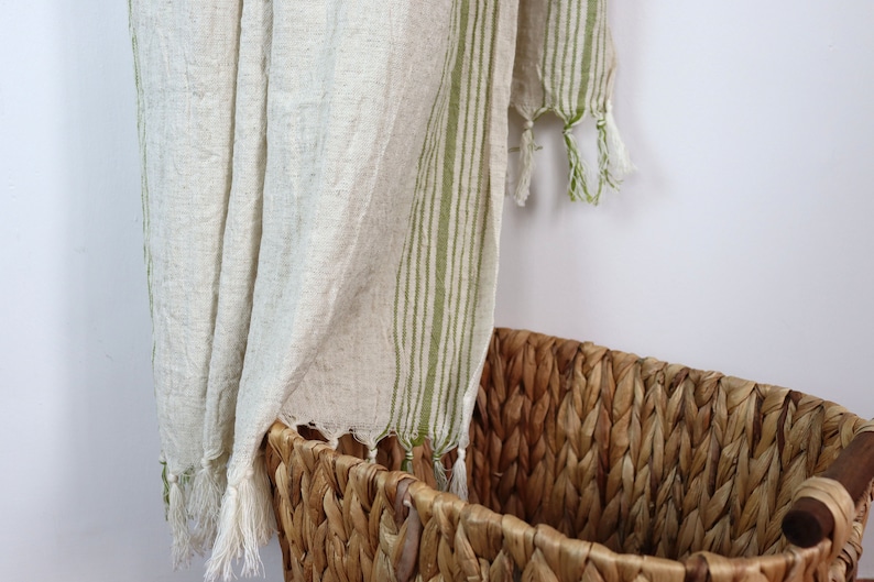 May include: A white linen throw with green stripes and tassels hanging over a woven wicker basket.
