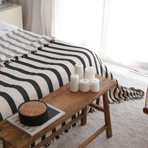 May include: A wooden bench with a black and white striped throw blanket on top. There are five white candles on the bench, and a woven basket with a black lid is on the bench.