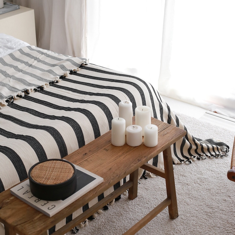 May include: A wooden bench with a black and white striped throw blanket on top. There are five white candles on the bench, and a woven basket with a black lid is on the bench.