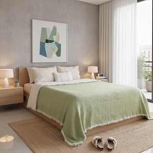 May include: A bedroom with a bed dressed in light green textured throw and white bedding. The room features a neutral rug, wooden bedside tables with lamps, and an abstract artwork. The decor is modern and minimalist, with a focus on natural light and textures.