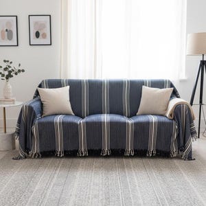 May include: A blue sofa draped with a striped throw, featuring vertical white and beige stripes, and two beige cushions. The throw has a fringed edge. The room is bright, with a patterned rug and a neutral colour scheme.