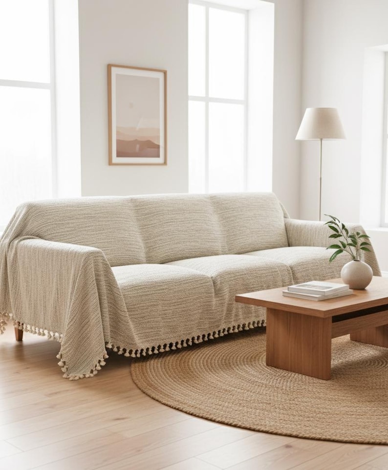 May include: A three-seater sofa covered in a textured, light beige fabric throw blanket. The blanket features a woven pattern and is finished with small, decorative tassels along the bottom edge, draping over the arms and seat of the sofa.