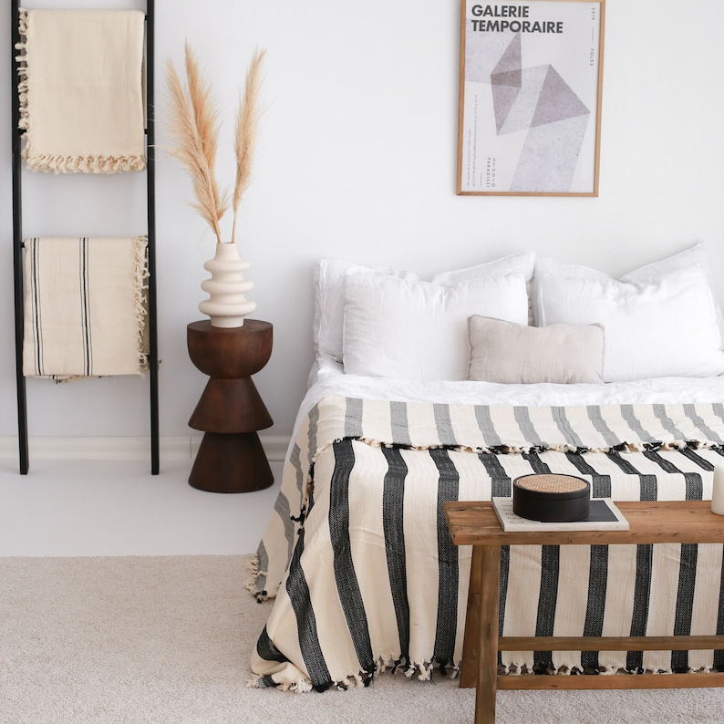 May include: A black and white striped throw blanket with fringe edges draped over a bed with white bedding. A wooden stool with a white vase and dried pampas grass sits next to the bed. A framed poster with the text "GALERIE TEMPORAIRE" hangs on the wall above the bed.