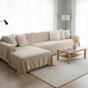 Könnte beinhalten: Beiges Ecksofa mit passenden Überwürfen und Kissen. Ein Couchtisch aus hellem Holz mit einem unteren Regal steht auf einem neutralen Teppich. Das Sofa ist mit einer strukturierten Decke mit Quastenbesatz bedeckt.