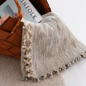 May include: A white and gray throw blanket with a fringe border. The blanket is folded and placed in a brown wicker basket.