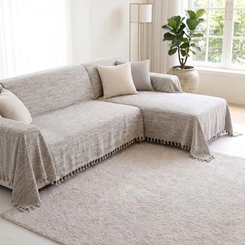 May include: A sectional sofa covered in a textured light gray fabric throw with fringe detailing along the bottom edges. The sofa features two solid cream-colored square throw pillows and one solid gray square throw pillow placed on the cushions.