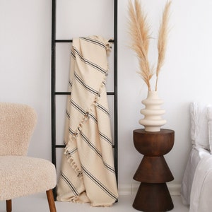 May include: A cream-colored throw blanket with black stripes hangs on a black metal ladder. The blanket has a fringe border. A vase with dried pampas grass sits on a wooden side table.