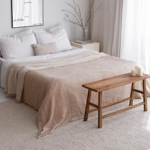 May include: A light brown throw blanket with a fringed edge draped over a bed with white linens. A wooden bench with a natural finish sits in front of the bed.