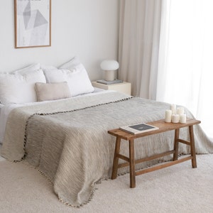 May include: A white bed with a gray and white striped throw blanket and a wooden bench with a book on top. The bed has white pillows and a beige throw pillow. There is a white lamp on a small table next to the bed.