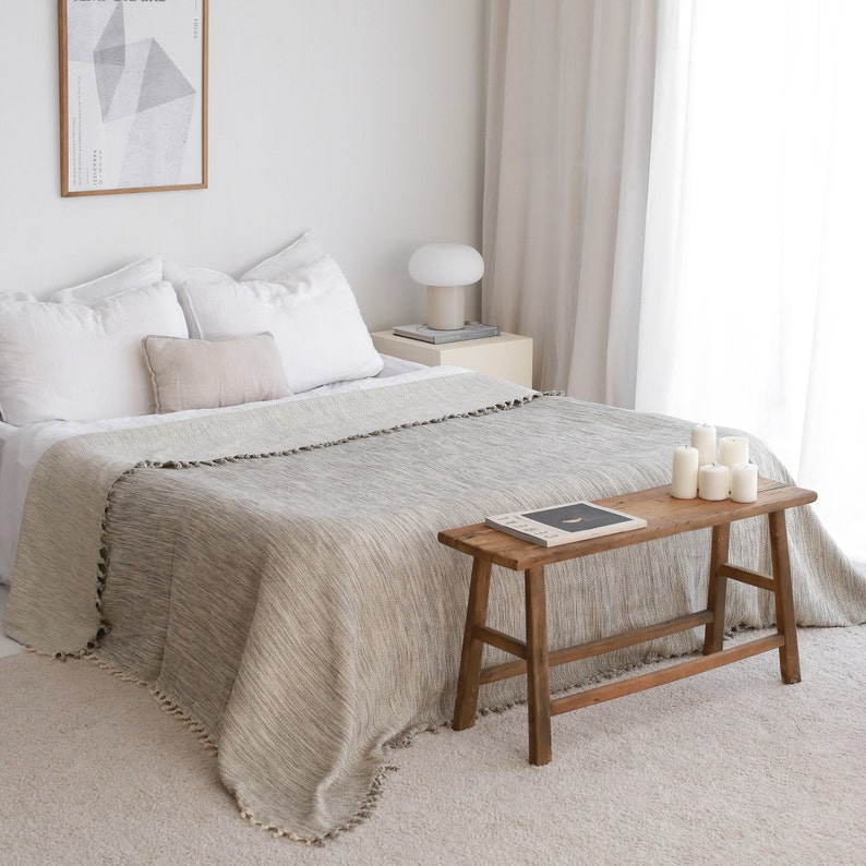 May include: A white bed with a gray and white striped throw blanket and a wooden bench with a book on top. The bed has white pillows and a beige throw pillow. There is a white lamp on a small table next to the bed.