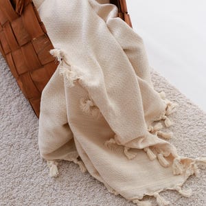 Puede incluir: Una cesta tejida marr&oacute;n contiene una manta color crema con flecos de borlas. La manta tiene un tejido texturizado y est&aacute; drapeada sobre el borde de la cesta. La cesta est&aacute; sobre una alfombra de color claro.