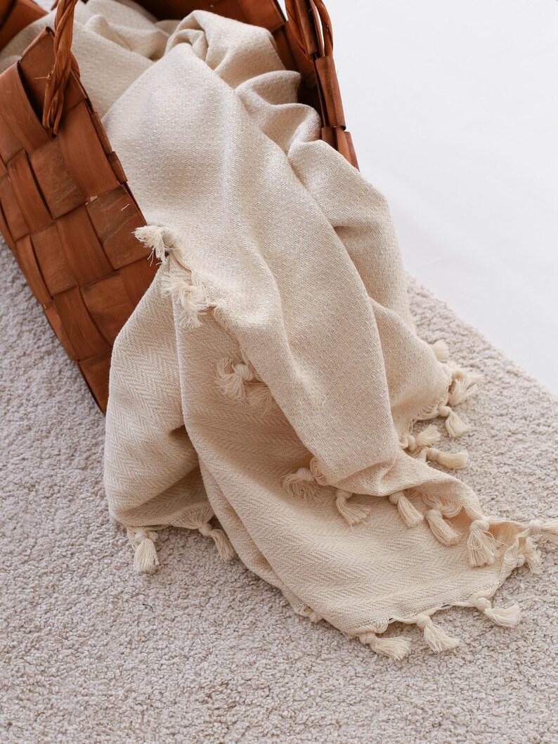 Puede incluir: Una cesta tejida marr&oacute;n contiene una manta color crema con flecos de borlas. La manta tiene un tejido texturizado y est&aacute; drapeada sobre el borde de la cesta. La cesta est&aacute; sobre una alfombra de color claro.