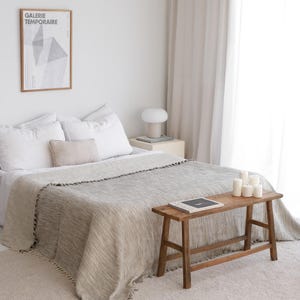 May include: A bedroom featuring a bed with white pillows and a beige textured throw blanket with fringe edges. A wooden bench sits at the foot of the bed, holding a book and white candles. A framed poster reads GALERIE TEMPORAIRE.