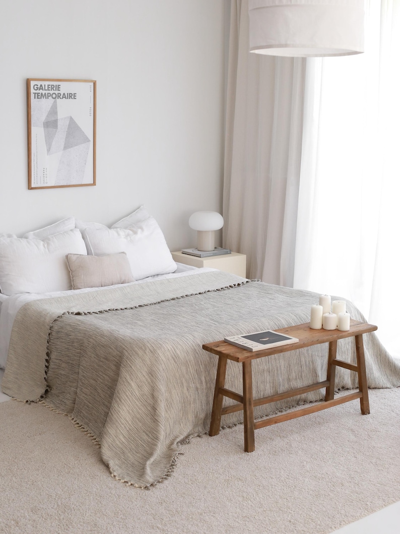 May include: A bedroom featuring a bed with white pillows and a beige textured throw blanket with fringe edges. A wooden bench sits at the foot of the bed, holding a book and white candles. A framed poster reads GALERIE TEMPORAIRE.