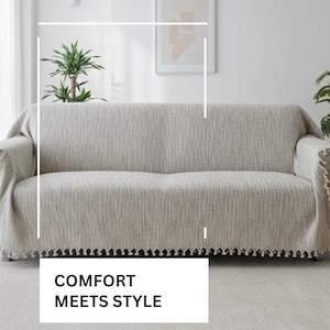 May include: A beige sofa cover with a textured, woven fabric finish and a decorative fringe along the bottom edge. The cover is draped over a sofa, featuring the text COMFORT MEETS STYLE in a white box, with additional text below the box.