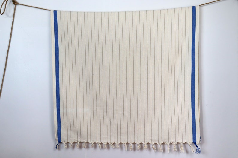 May include: A white throw blanket with thin brown stripes and blue borders. The blanket has a fringe on the bottom edge.