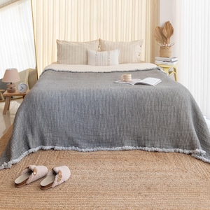 Puede incluir: Una manta de tejido de gofre gris con borde de flecos está drapeada sobre una cama con almohadas blancas y beige. La manta está sobre una alfombra de fibra natural.