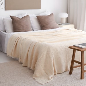 Puede incluir: Una manta de tiro de color crema con un tejido texturizado y bordes con flecos está drapeada sobre una cama con almohadas marrones. La manta está sobre una cama blanca con una cabecera blanca y una mesita de noche blanca con una lámpara.