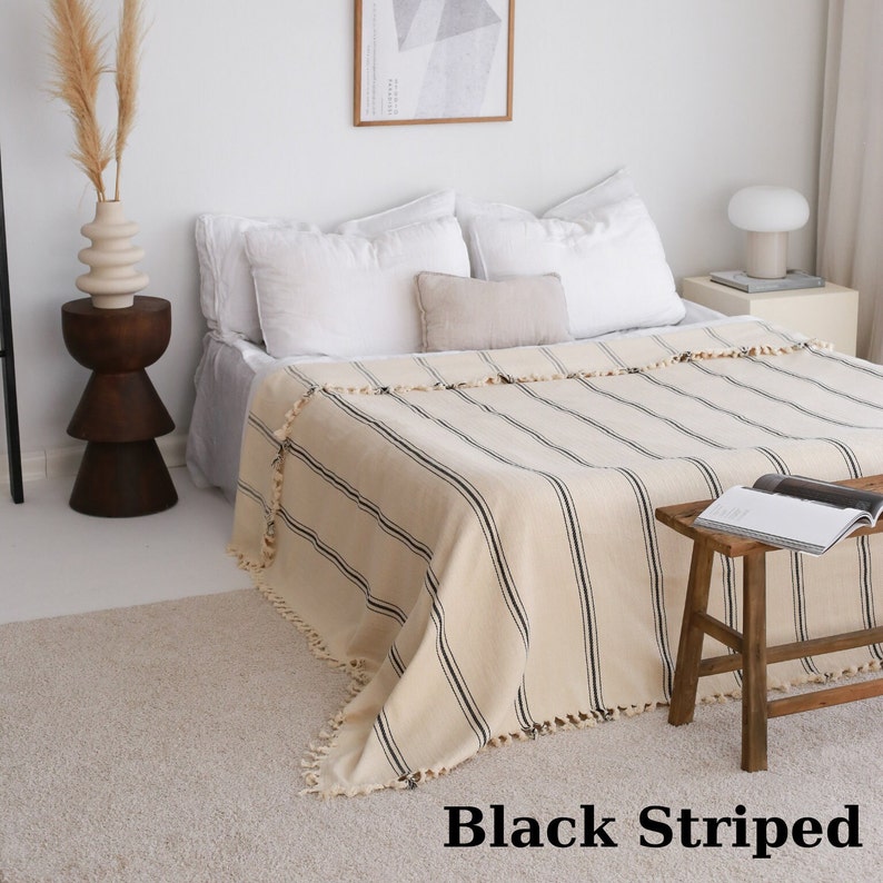 May include: A cream-colored throw blanket with black stripes and fringe trim. The blanket is draped over a bed with white pillows.
