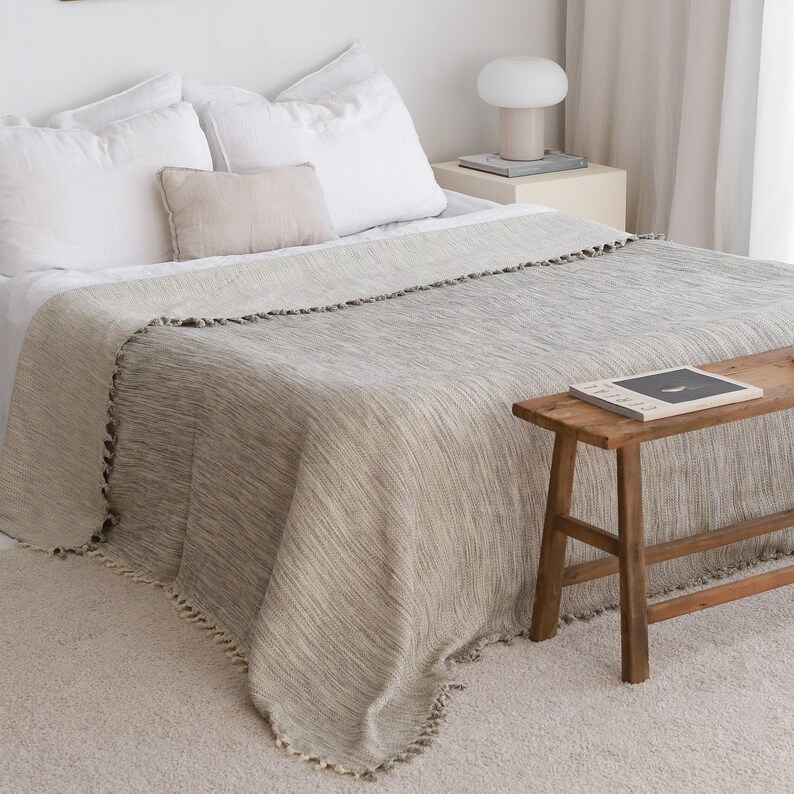 May include: A gray and white striped throw blanket with a fringe border. The blanket is draped over a bed with white pillows and a small wooden stool.