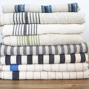 May include: A stack of eight striped linen towels in various colors, including black, white, blue, and green. The towels are folded and stacked neatly on a wooden surface.