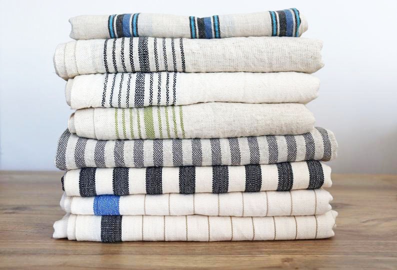 May include: A stack of eight striped linen towels in various colors, including black, white, blue, and green. The towels are folded and stacked neatly on a wooden surface.