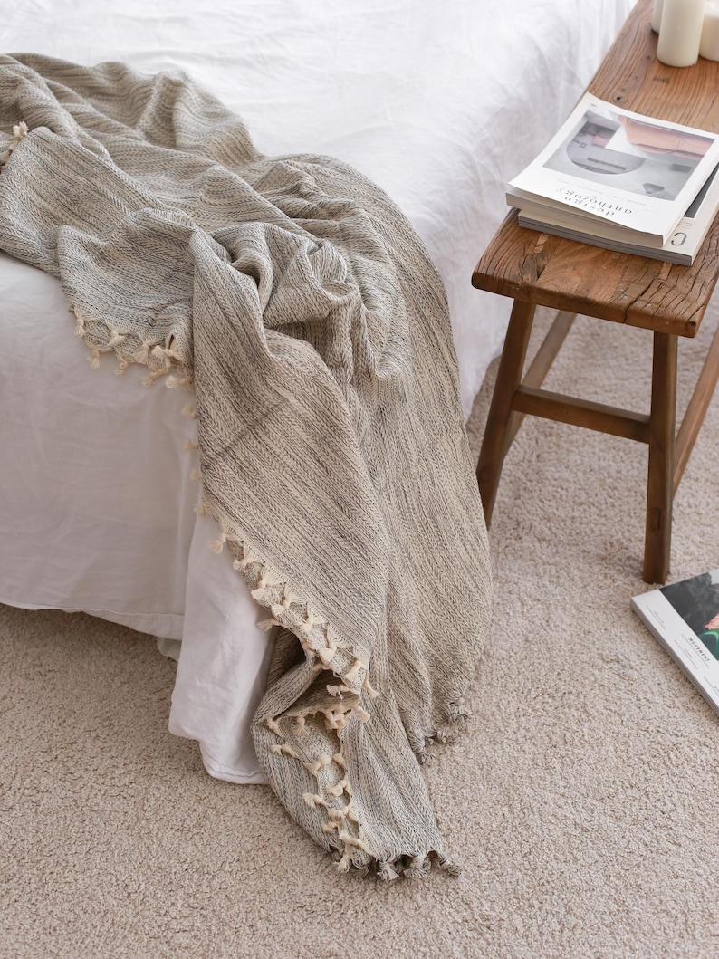 May include: A woven throw blanket featuring a textured pattern in shades of beige, gray, and white. The blanket has a decorative fringe trim along the edges with small, knotted tassels in a cream color, draped over a white bed sheet.