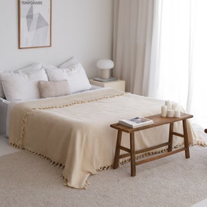 May include: A beige throw blanket with fringe trim is draped over a bed with white pillows. A wooden bench with a magazine on top sits in front of the bed.