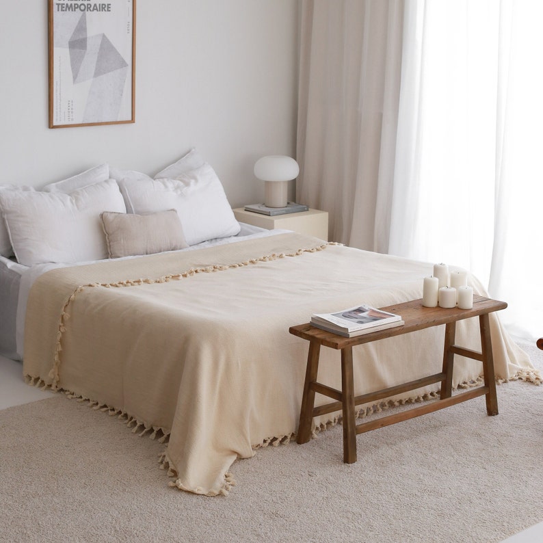May include: A beige throw blanket with fringe trim is draped over a bed with white pillows. A wooden bench with a magazine on top sits in front of the bed.