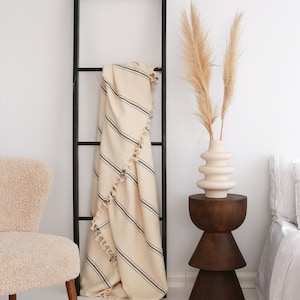 Puede incluir: Una escalera de madera negra se apoya en una pared blanca, con una manta crema con rayas negras y borlas. Una silla beige y una mesa auxiliar de madera con un jarr&oacute;n de pampas completan la escena, creando un ambiente acogedor.