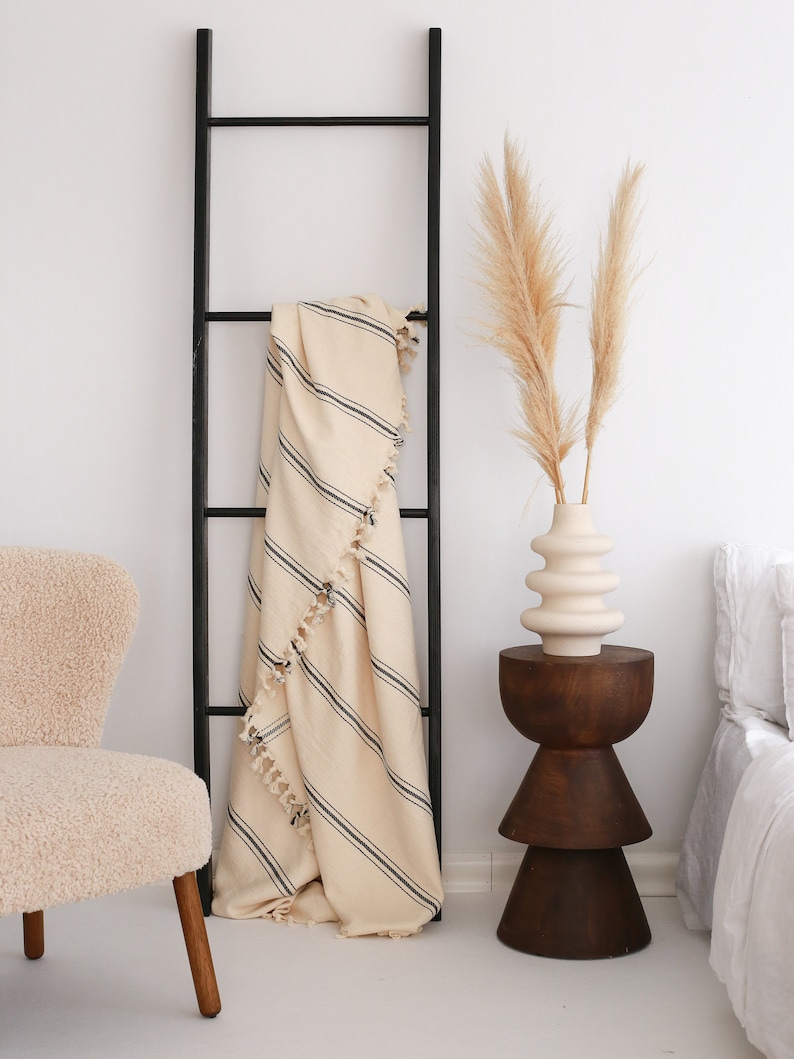 Puede incluir: Una escalera de madera negra se apoya en una pared blanca, con una manta crema con rayas negras y borlas. Una silla beige y una mesa auxiliar de madera con un jarr&oacute;n de pampas completan la escena, creando un ambiente acogedor.