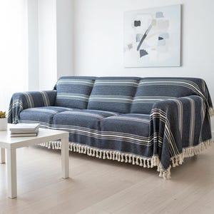 May include: A three-seat sofa covered in a dark blue textured fabric throw blanket featuring horizontal white stripes and a decorative fringe along the bottom edge. The blanket drapes over the back, seat, and arms of the sofa, providing full coverage.