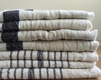 May include: A stack of folded linen towels. The towels are primarily off-white with black and brown stripes and accents. The towels are stacked neatly on a wooden surface, creating a clean and organized display.