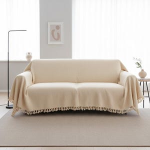 Puede incluir: Un sofá color crema está cubierto con una manta a juego. La manta tiene flecos a lo largo del borde inferior. El sofá se encuentra sobre una alfombra de color neutro en una habitación con cortinas blancas y una obra de arte abstracta enmarcada.