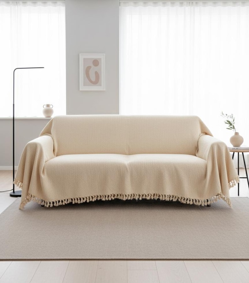 Puede incluir: Un sof&aacute; color crema est&aacute; cubierto con una manta a juego. La manta tiene flecos a lo largo del borde inferior. El sof&aacute; se encuentra sobre una alfombra de color neutro en una habitaci&oacute;n con cortinas blancas y una obra de arte abstracta enmarcada.