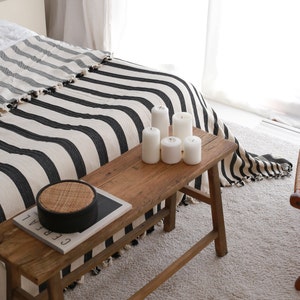 May include: A wooden bench with a black and white striped throw blanket draped over it. The bench has a magazine and a woven round tray on top. There are five white candles on the bench.