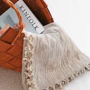 May include: A woven brown basket containing a Kinfolk magazine and a light gray and white textured throw blanket with small cream-colored tassels along the edges. The blanket features a subtle herringbone pattern and hangs over the basket rim.