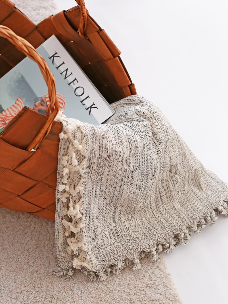 May include: A woven brown basket containing a Kinfolk magazine and a light gray and white textured throw blanket with small cream-colored tassels along the edges. The blanket features a subtle herringbone pattern and hangs over the basket rim.