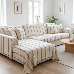 May include: A cream-coloured sectional sofa with a navy blue striped pattern. The sofa is draped with matching throws and cushions. The throws have a fringe trim. The room has a neutral colour palette with light-coloured walls and a wooden coffee table.