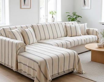 May include: A large sectional sofa covered in a cream-colored fabric with thin vertical black stripes. The sofa features a matching throw blanket draped over the chaise section, several square throw pillows, and decorative fringe along the edges.