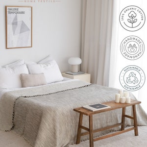 May include: A light gray throw blanket with fringe trim is draped over a bed with white linens. A wooden stool with a book on top sits in front of the bed. The room has a white wall with a framed print and a white lamp on a side table. The image includes the text "Budapestmal Home Textile" and several logos indicating the blanket is made of 100% organic cotton, is handmade, and is sustainable.
