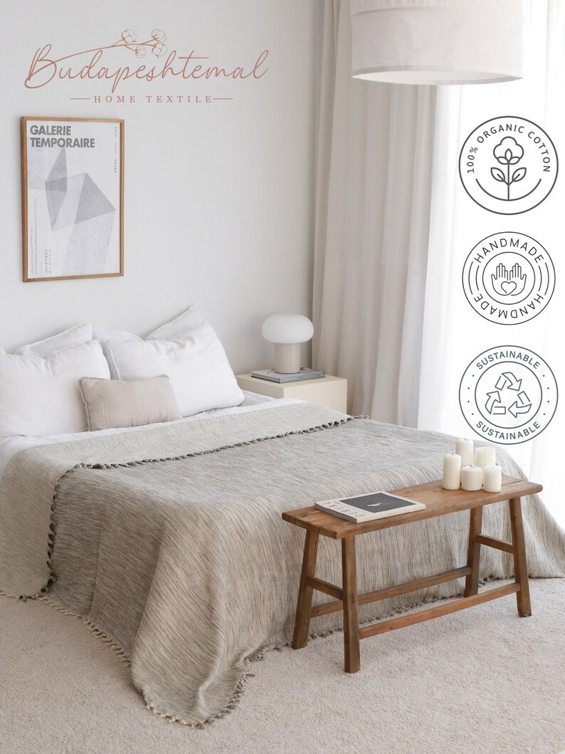 May include: A light gray throw blanket with fringe trim is draped over a bed with white linens. A wooden stool with a book on top sits in front of the bed. The room has a white wall with a framed print and a white lamp on a side table. The image includes the text "Budapestmal Home Textile" and several logos indicating the blanket is made of 100% organic cotton, is handmade, and is sustainable.