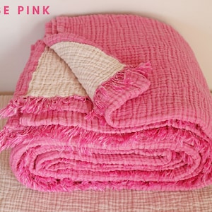 Pode incluir: Uma manta de algodão rosa com um lado inferior branco e bordas franjadas. A manta está enrolada e tem um aspeto suave e texturizado.