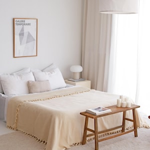 Puede incluir: Una habitaci&oacute;n con una cama cubierta con ropa de cama blanca y una manta beige con flecos. Un banco de madera sostiene velas y un libro. Un p&oacute;ster enmarcado con el texto "GALERIE TEMPORAIRE" cuelga de la pared.