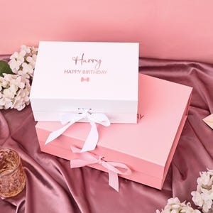 Puede incluir: Dos cajas de regalo, una blanca y otra rosa, apiladas sobre una superficie de satén. La caja blanca tiene el nombre "Harry" y "HAPPY BIRTHDAY" impresos en la tapa, con un lazo gráfico. Ambas cajas están atadas con cintas.