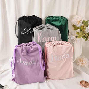 Puede incluir: Cinco bolsas de terciopelo con cordón en negro, gris, verde, lavanda y rosa. Cada bolsa tiene un nombre bordado en un color contrastante: "He", "Karen", "Jessica" y "Linda". Las bolsas están dispuestas sobre una superficie blanca.