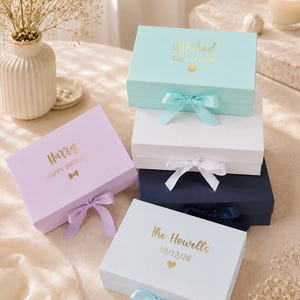 Puede incluir: Una pila de cinco cajas de regalo rectangulares en azul claro, blanco, azul marino y lavanda, cada una con un cierre de cinta de satén. Las cajas están personalizadas con texto en lámina dorada que incluye nombres como Michael, Harry y The Howells, y fechas.