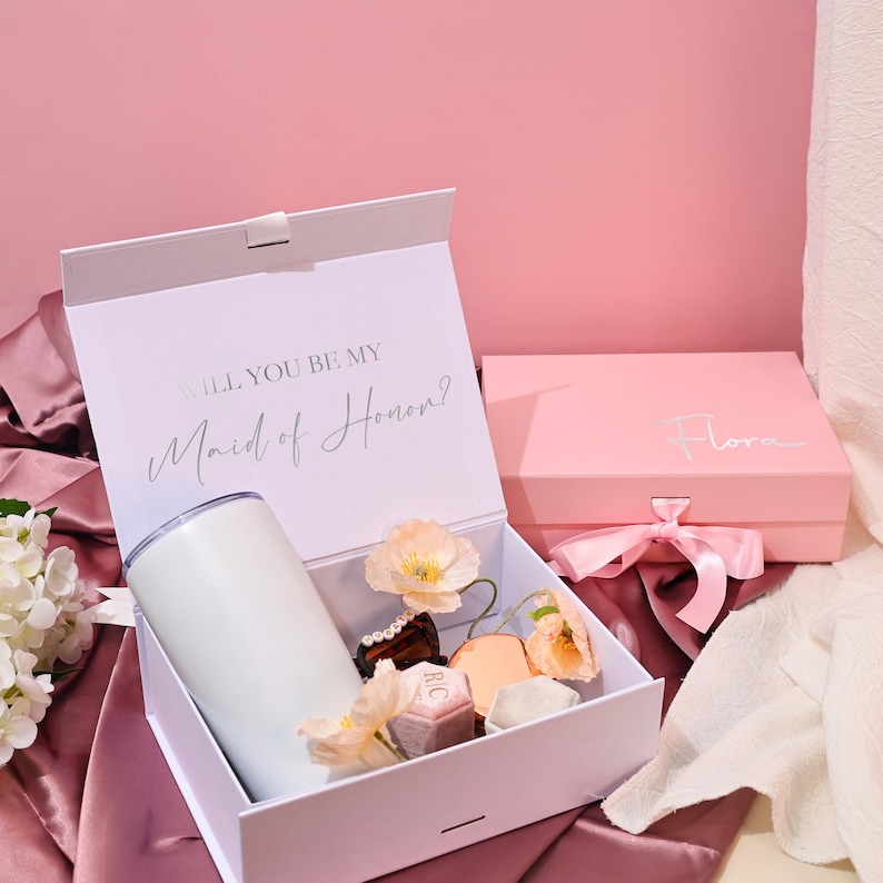 May include: A white gift box with the text "WILL YOU BE MY Maid of Honor?" contains a white tumbler, small gifts, and flowers. A pink box with a bow and the name "Flora" is also present. The background is pink.